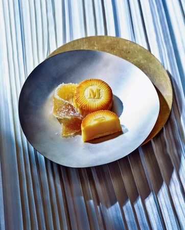 Elegant food still life featuring cut mooncakes with an 'M' logo, alongside candied fruit, served on silver and gold plates.