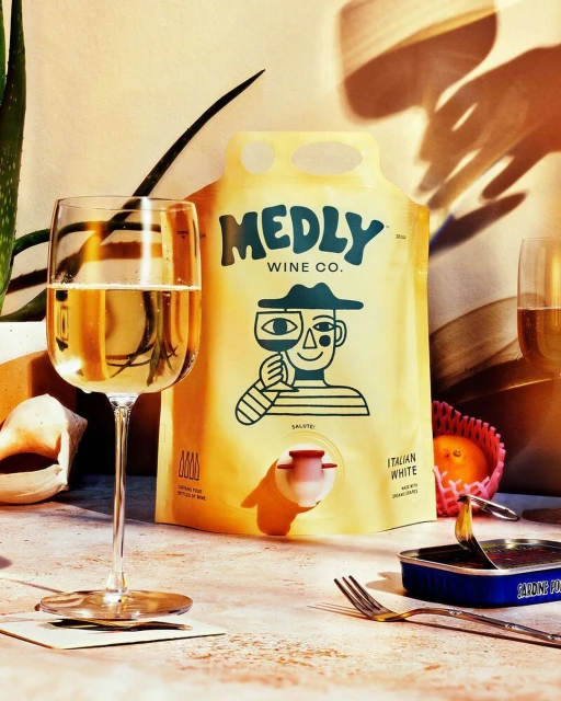 Still life photo of a Medly Italian White wine bag-in-box with a wine glass, sardines, fruit, and plant against a beige wall.