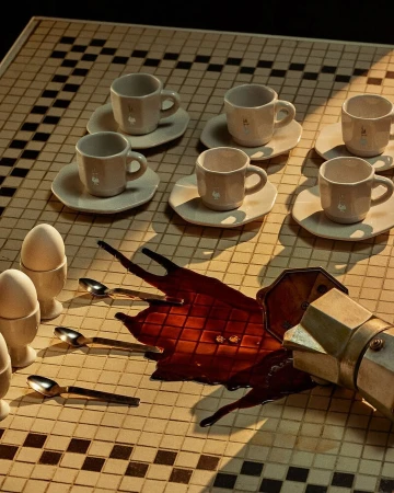 Still life with spilled coffee, espresso maker, cups, eggs, and spoons on a checkered tile surface with studio lighting.