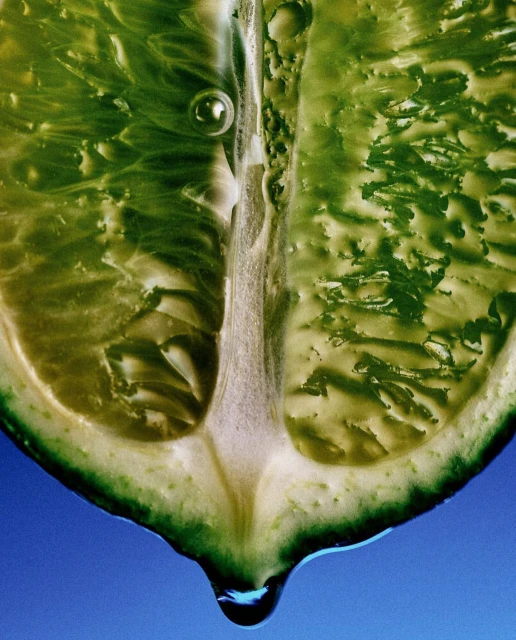 Macro close-up of a sliced lime dripping water, showcasing its vibrant green color and juicy texture against a blue background.