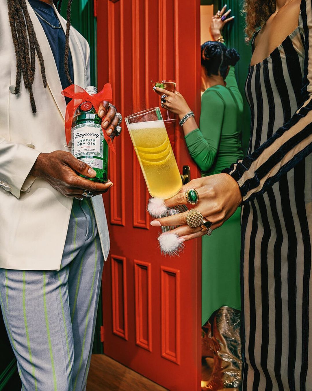 Three people with drinks pose indoors against a red and green wall. The photo features a cocktail and a gin bottle.
