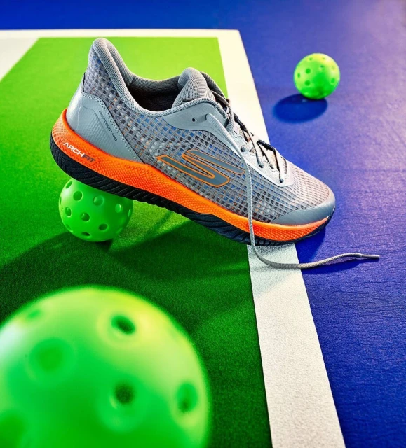 Gray Skechers sneaker with orange sole balancing on a green pickleball on a blue and green pickleball court with green pickleballs.
