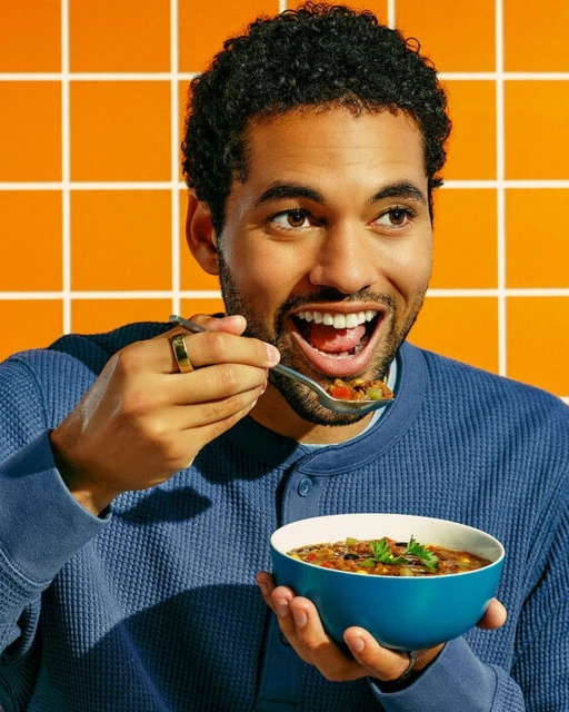 Man eating soup from a blue bowl. The man smiles with open mouth in an indoor setting with an orange, tiled background.
