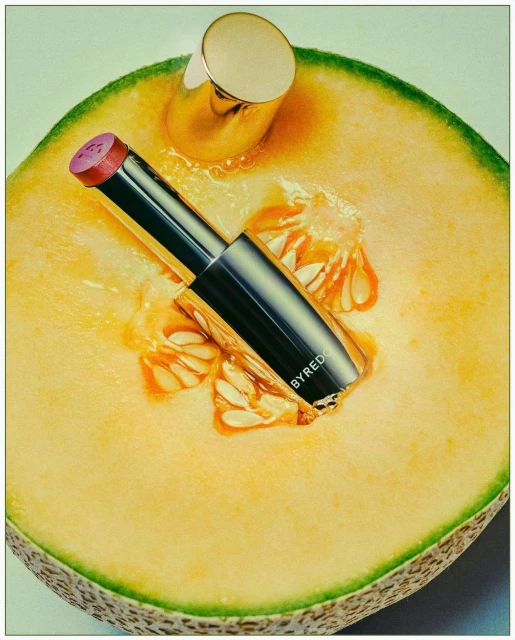 Still life of a tube of BYREDO lipstick displayed on a cantaloupe melon half, showcasing makeup and food in a close-up shot.
