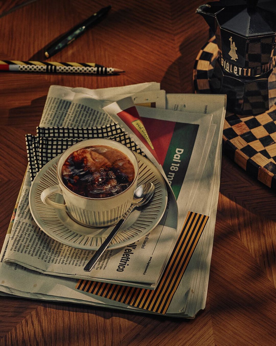 A cup of dark coffee sits on a stack of newspapers next to pens and a stovetop espresso maker on a wood table.