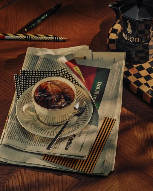 A cup of dark coffee sits on a stack of newspapers next to pens and a stovetop espresso maker on a wood table.