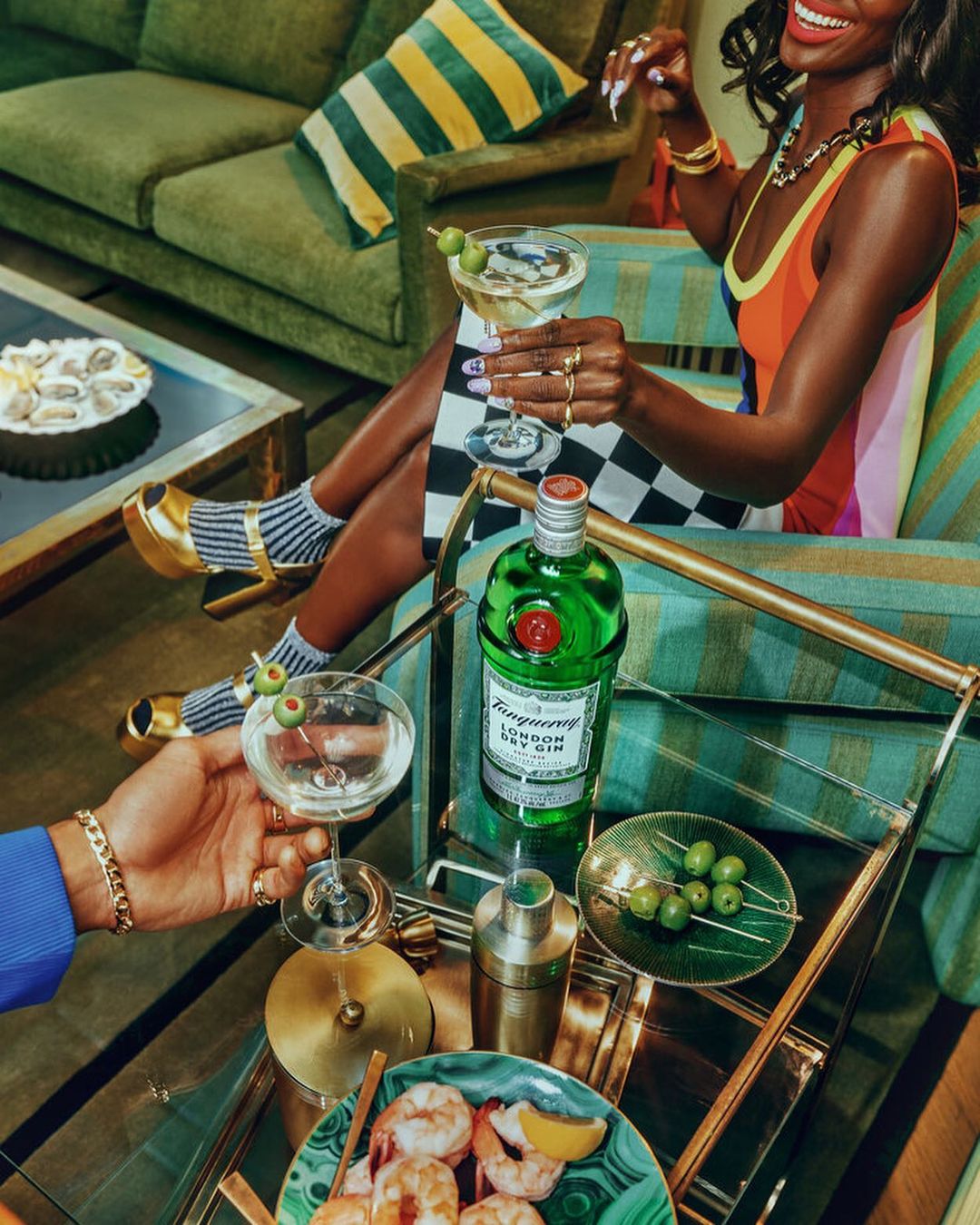 A smiling Black woman sits in a vintage armchair, holding a cocktail. Gin bottle, olives, and shrimp cocktail on a glass serving cart.