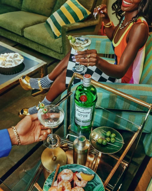 A smiling Black woman sits in a vintage armchair, holding a cocktail. Gin bottle, olives, and shrimp cocktail on a glass serving cart.