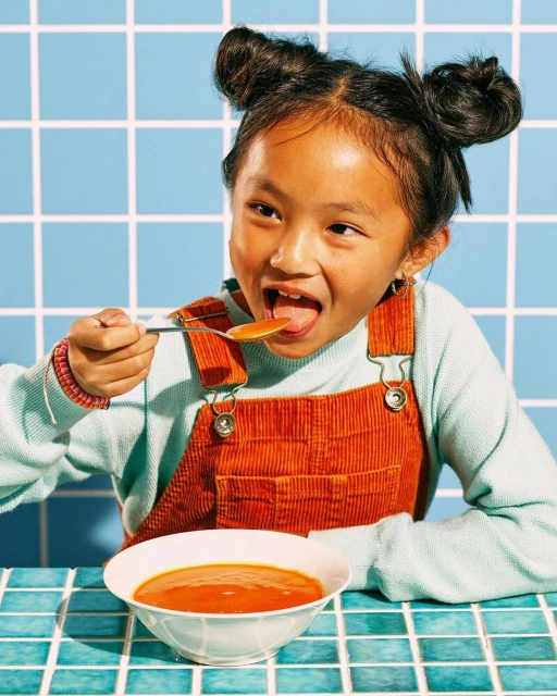 Young girl eating soup in a retro-tiled kitchen. The girl has an open mouth and a spoon near her face. Focus on a bowl of tomato soup.