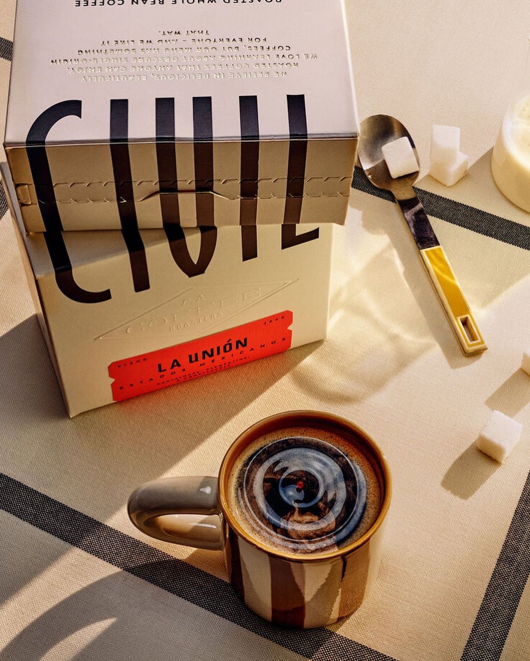 Coffee mug filled with black coffee, coffee boxes, sugar cubes, and a spoon arranged on a striped surface in a still life.