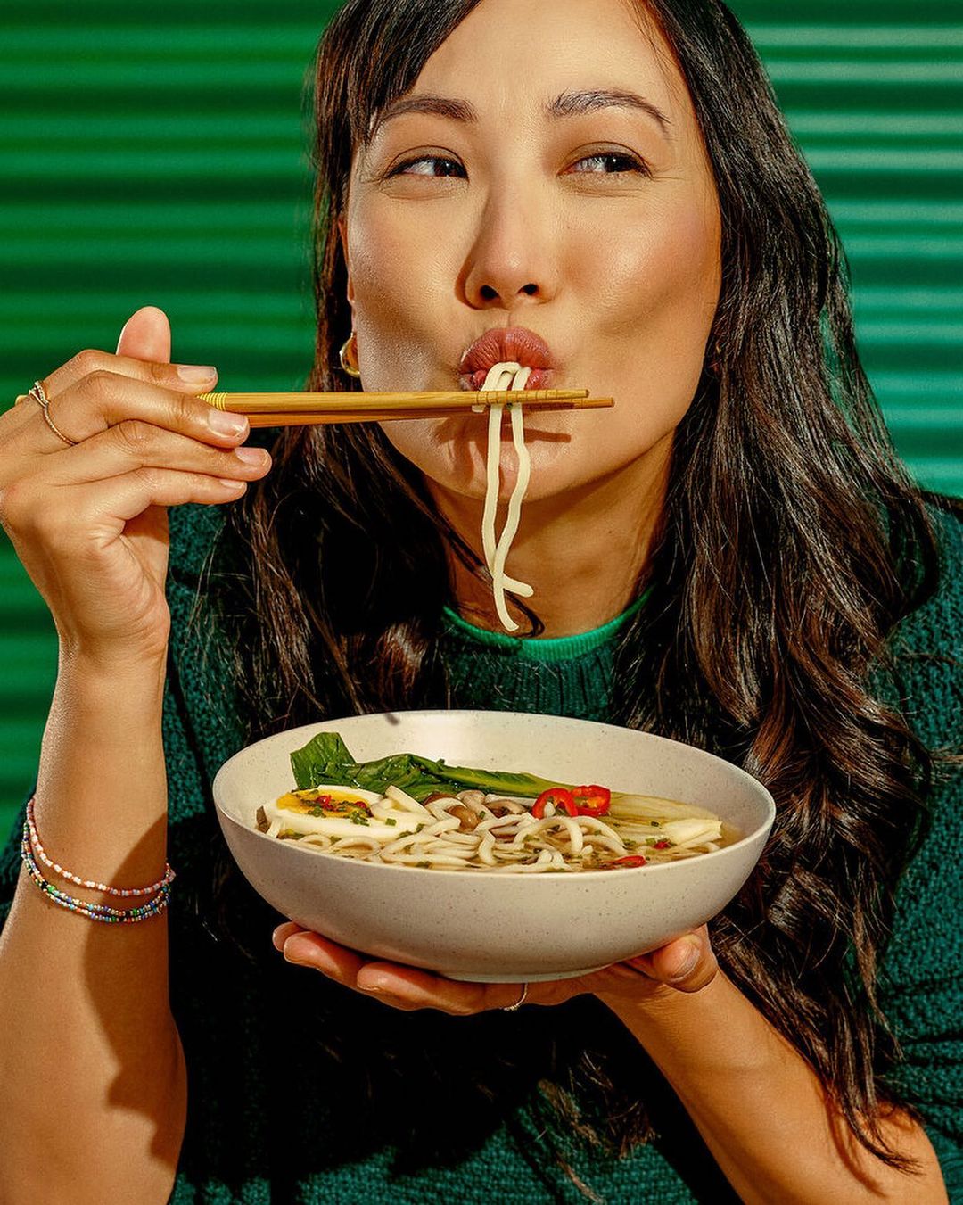 Asian woman enjoys noodles with chopsticks in a green-toned studio, highlighting the food's textures and her dining experience.