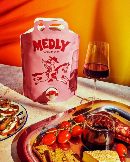 A wine bag and glass of red wine sit on a table with sliced bread, tomatoes, and cheese in a bright, eye-level product shot.