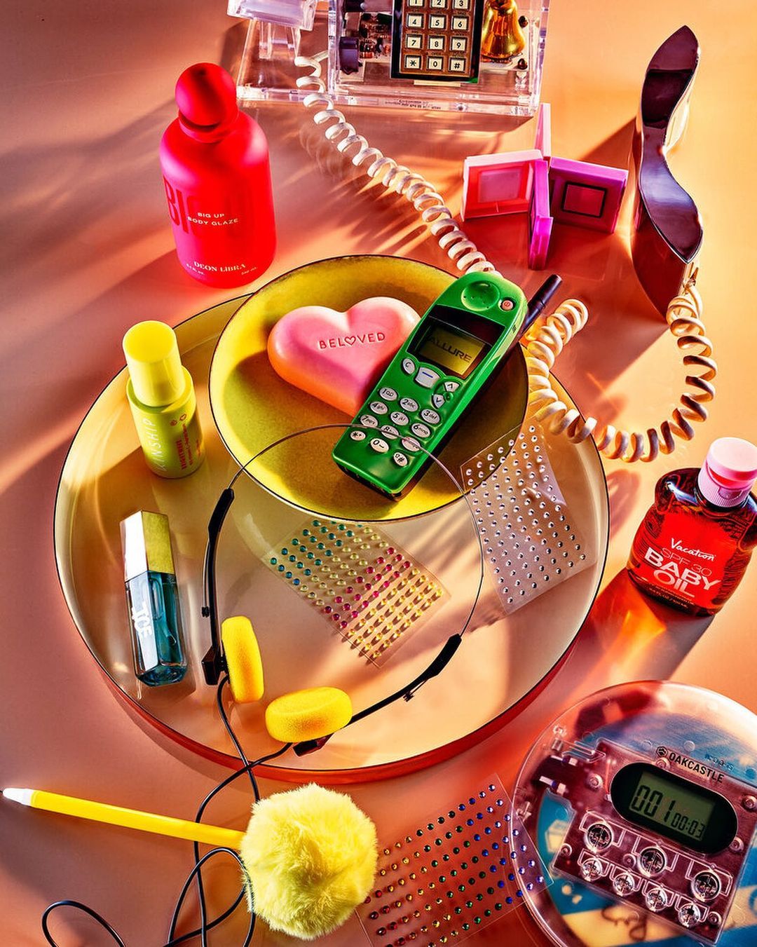 Still life of retro accessories and beauty products arranged on a round plate, featuring headphones, a toy phone, and skincare items.