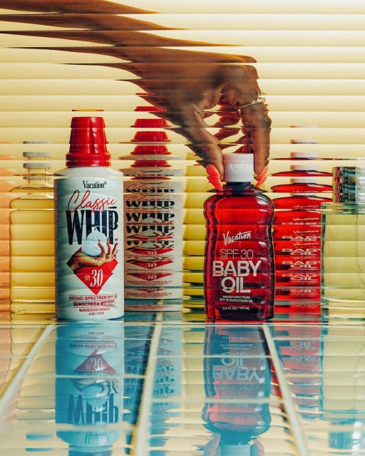 Various skincare and sunscreen products from Vacation brand with a hand in the frame on a glossy surface in front of blinds.