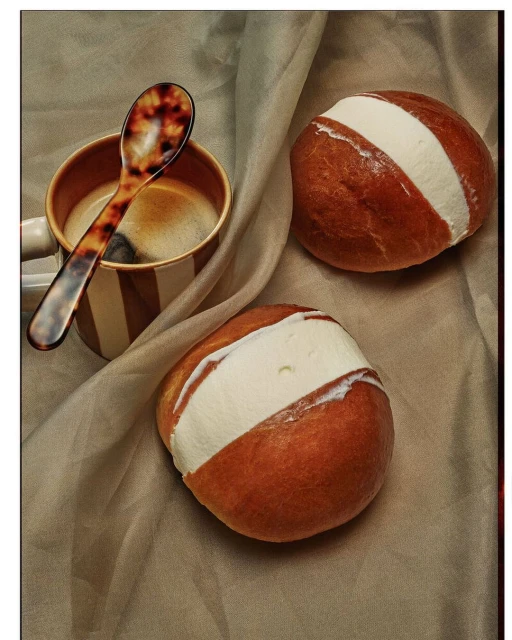 Two Sicilian brioche buns filled with gelato, accompanied by a spoon and a striped coffee mug, all resting on a draped fabric.