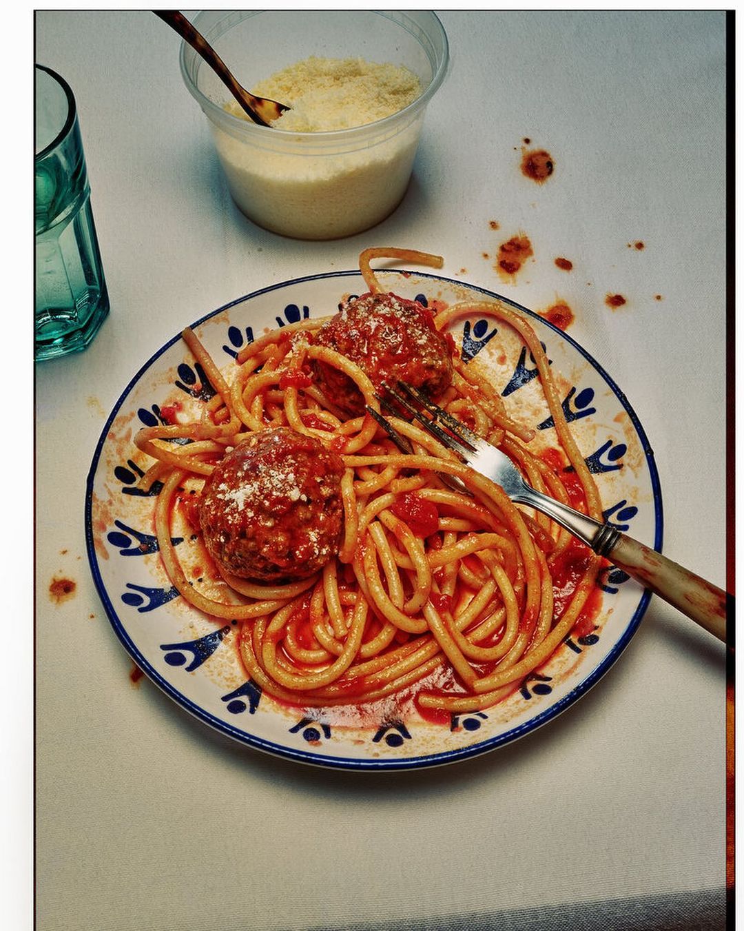 Overhead shot of pasta with tomato sauce and meatballs on a patterned plate next to a glass of water and parmesan.