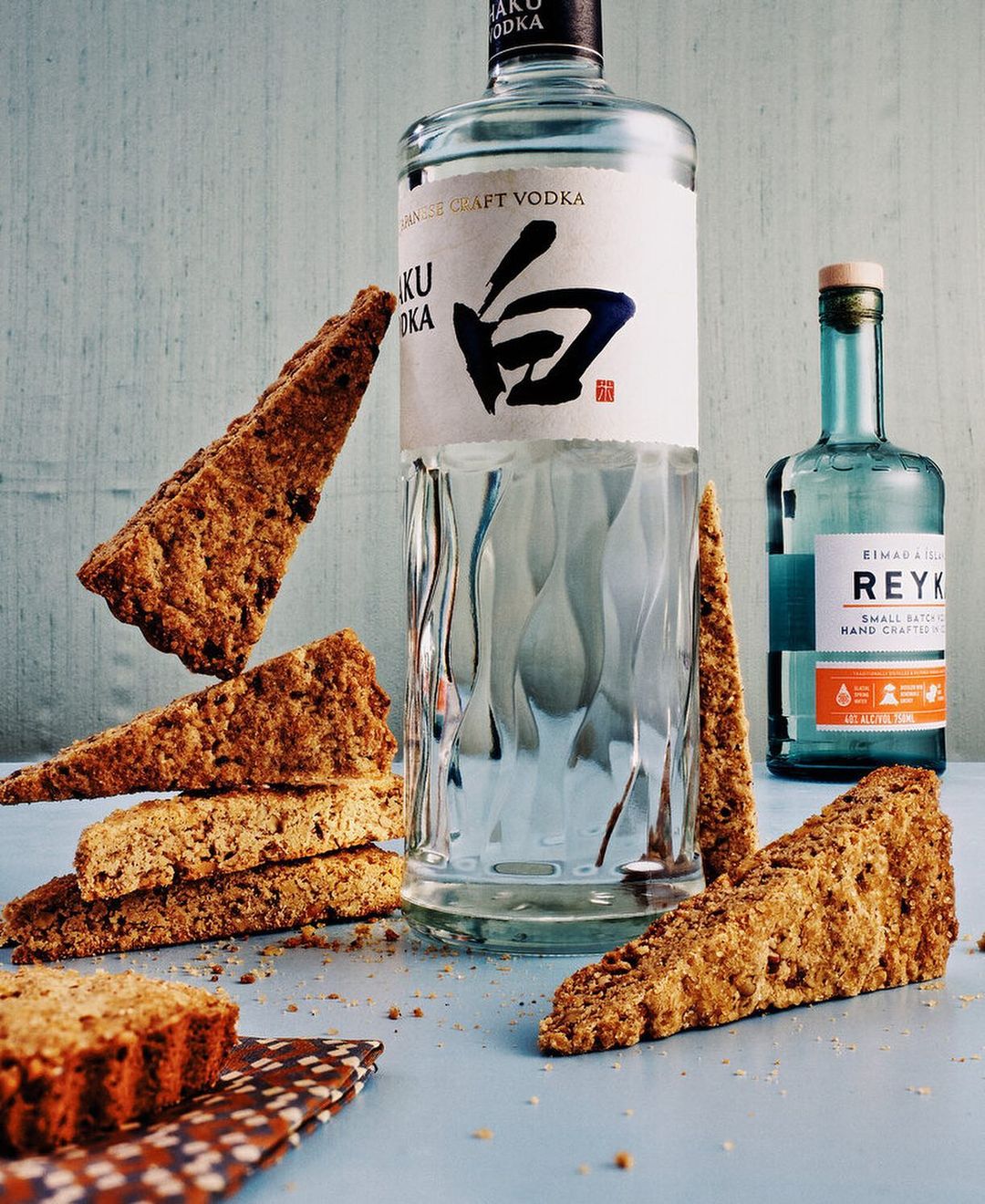 Still life shot of Haku vodka, Reyka vodka, and biscotti cookies on a blue surface with a light blue wall in the background.
