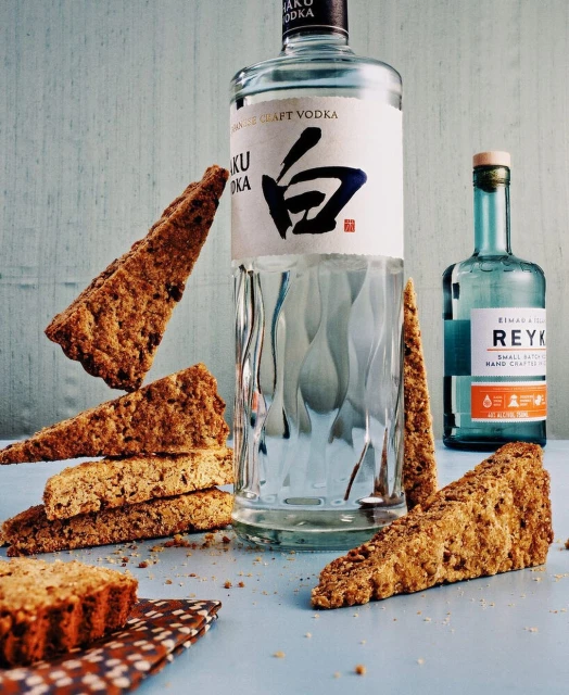 Still life shot of Haku vodka, Reyka vodka, and biscotti cookies on a blue surface with a light blue wall in the background.
