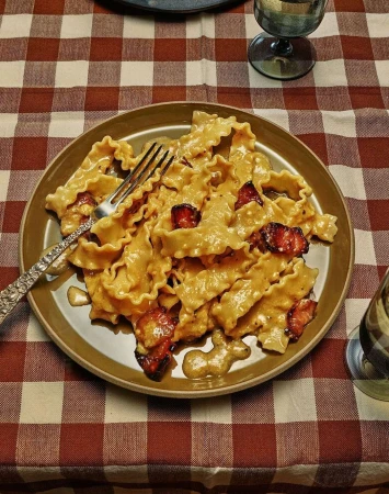 Close-up of pasta dish with a creamy sauce and meat, plated on a brown plate with a silver fork, set on a red and white checkered tablecloth.