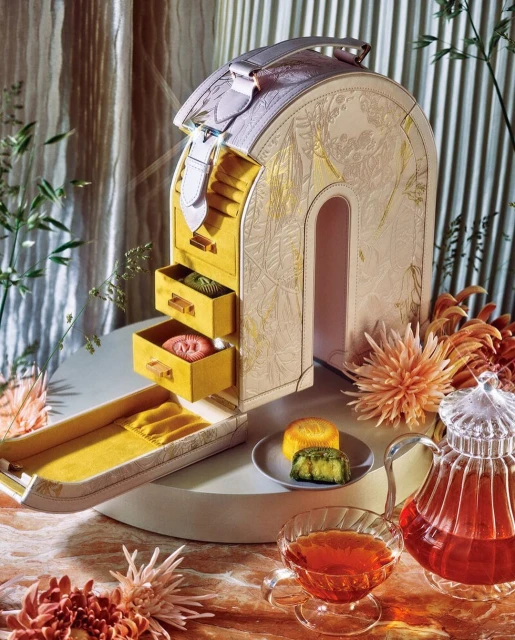 Luxurious gift box resembling a cabinet with mooncakes on a plate and a tea set, set against a backdrop of golden drapes and foliage.