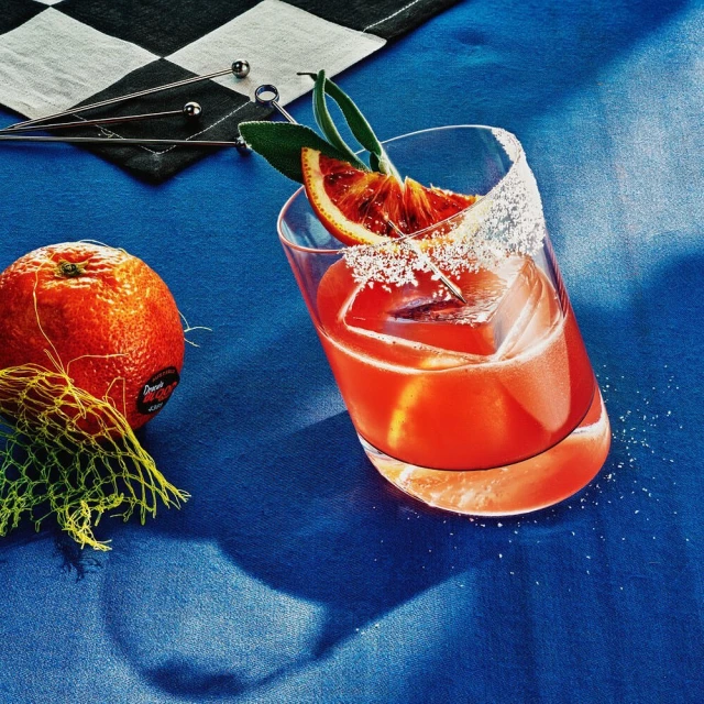 A vibrant still life showcases a cocktail in a sugar-rimmed glass garnished with an orange slice, alongside a whole orange and a checkered cloth.