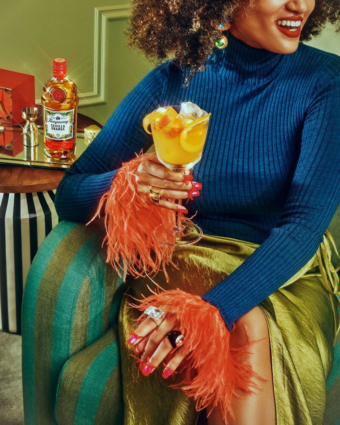Woman in feathered sleeves sips cocktail; liquor bottle & silver jigger on tray, studio lighting, food and beverage photography.