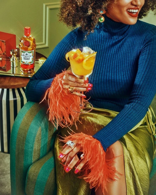 Woman in feathered sleeves sips cocktail; liquor bottle & silver jigger on tray, studio lighting, food and beverage photography.
