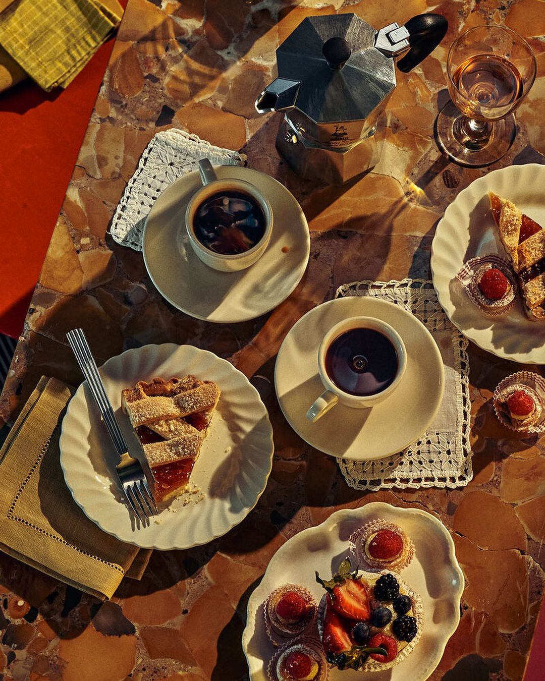 Overhead shot of coffee, pastries, tarts, and a coffee maker on a patterned marble table with sunlight creating shadows.