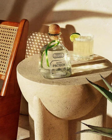 Patron tequila bottle with lime garnish and a cocktail on a round beige table with a cane chair and agave plant nearby.