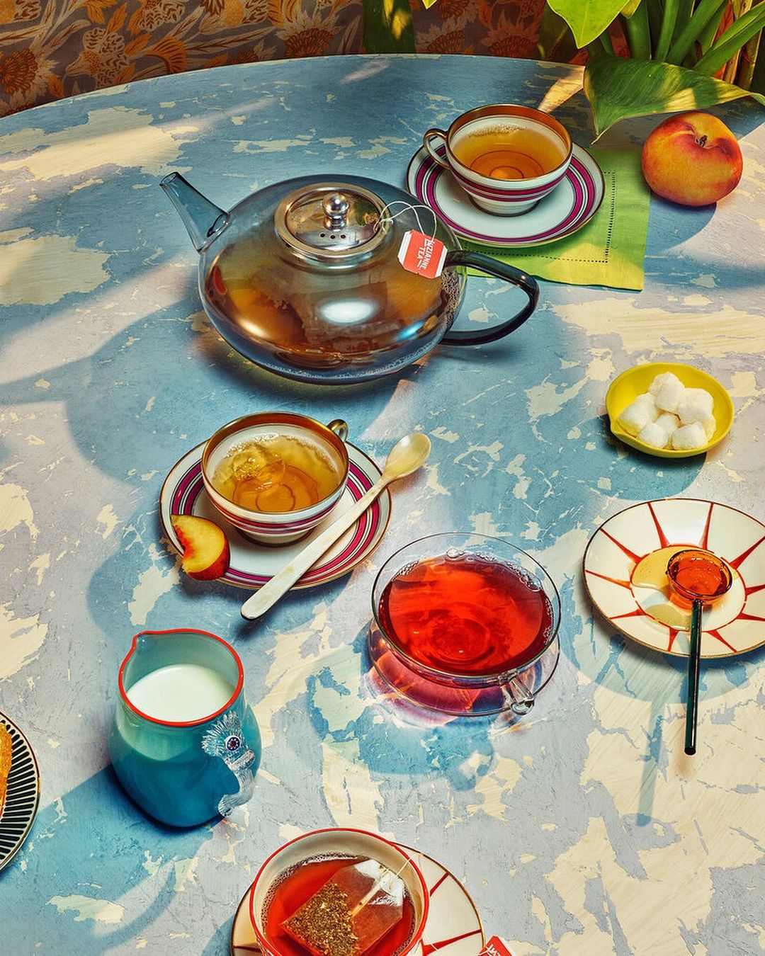 Overhead shot showcases a clear glass teapot, teacups with tea, a milk pitcher, sugar cubes, spoons, and a peach on a patterned tablecloth.