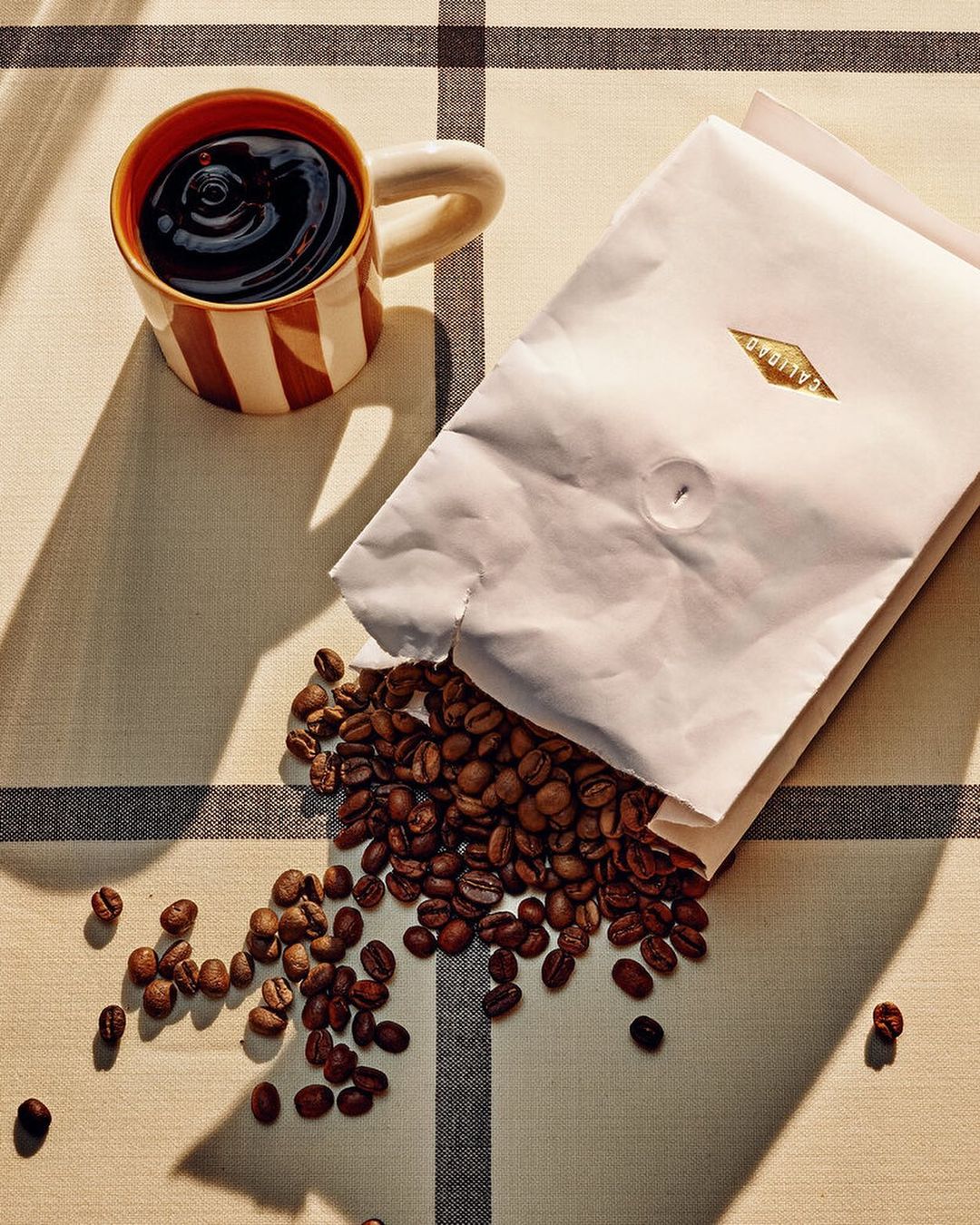 Still life shot of coffee beans spilling from a white bag next to a striped mug filled with coffee, all on a tan checkered tablecloth.