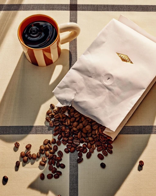 Still life shot of coffee beans spilling from a white bag next to a striped mug filled with coffee, all on a tan checkered tablecloth.