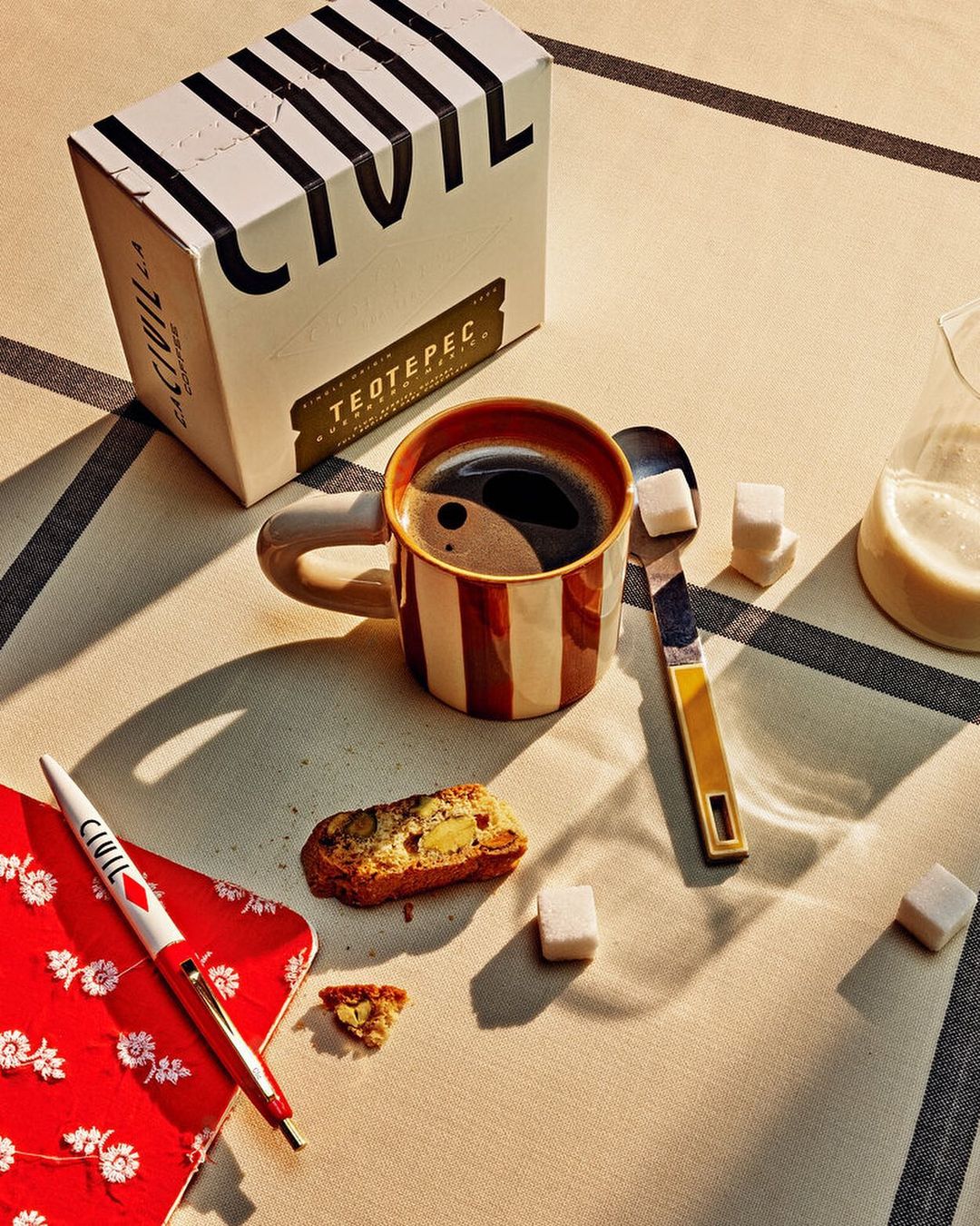 Still life of coffee cup, biscotti, and sugar cubes on a striped tablecloth with a red patterned notebook and pen, and coffee box.