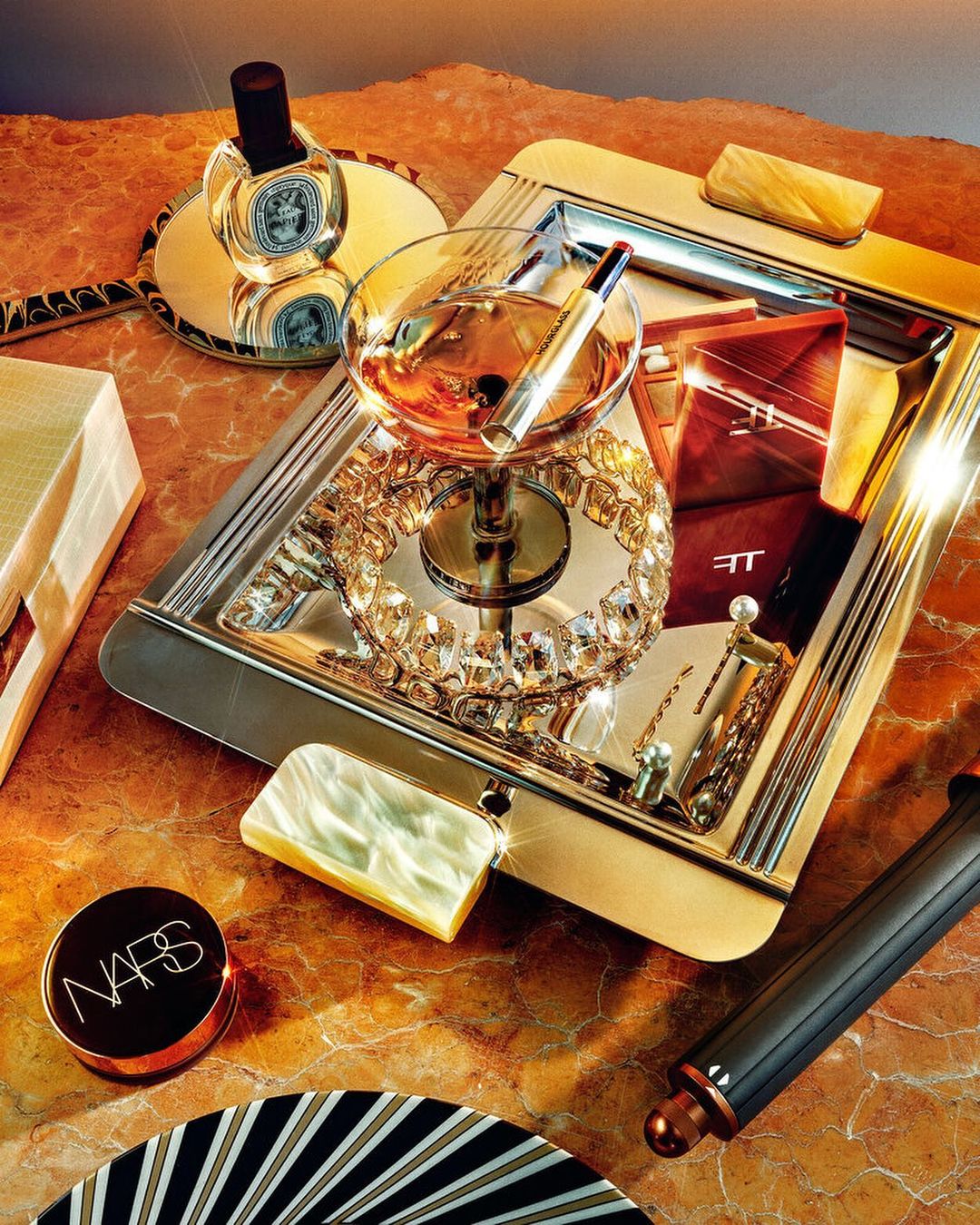 Still life photograph of cosmetics and beauty products, including a perfume bottle, lipstick, and compacts on a reflective surface.