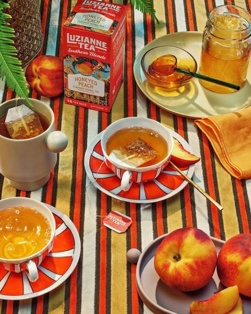A still life features cups of Luzianne Honeyed Peach tea, peaches, honey jar, and striped tablecloth, creating a warm and inviting scene.