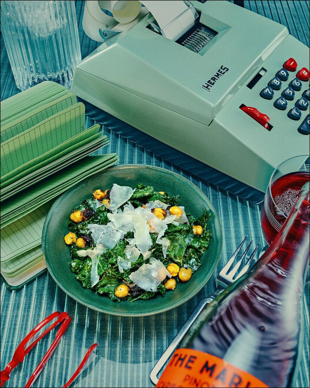 Overhead view of a kale salad with parmesan, a bottle of red wine, an adding machine, and notebooks on a teal striped surface.