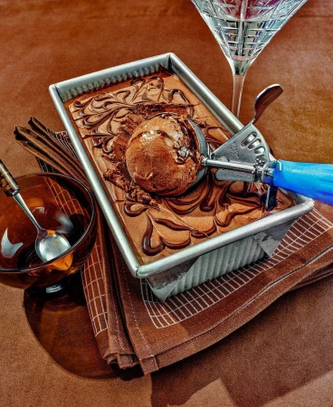 Chocolate ice cream scoop on top of homemade brownies in a metal baking pan with a glass bowl and cocktail glass on a brown surface.