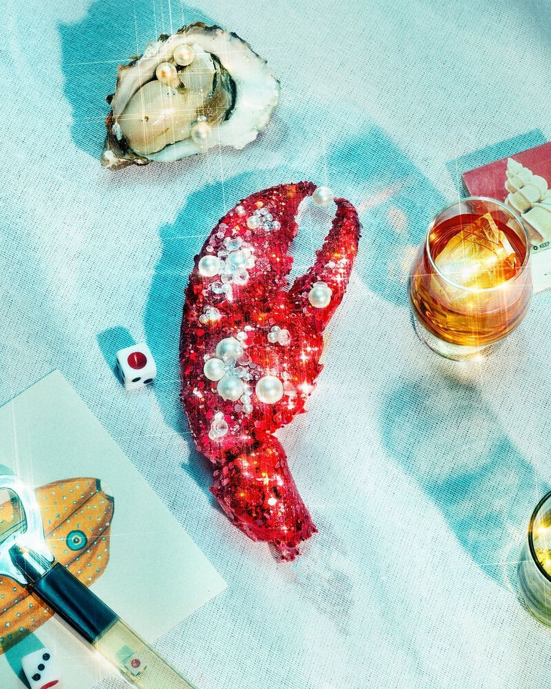Still life with a lobster claw and oyster adorned with pearls, a glass of amber liquid, and a dice on a textured white surface.