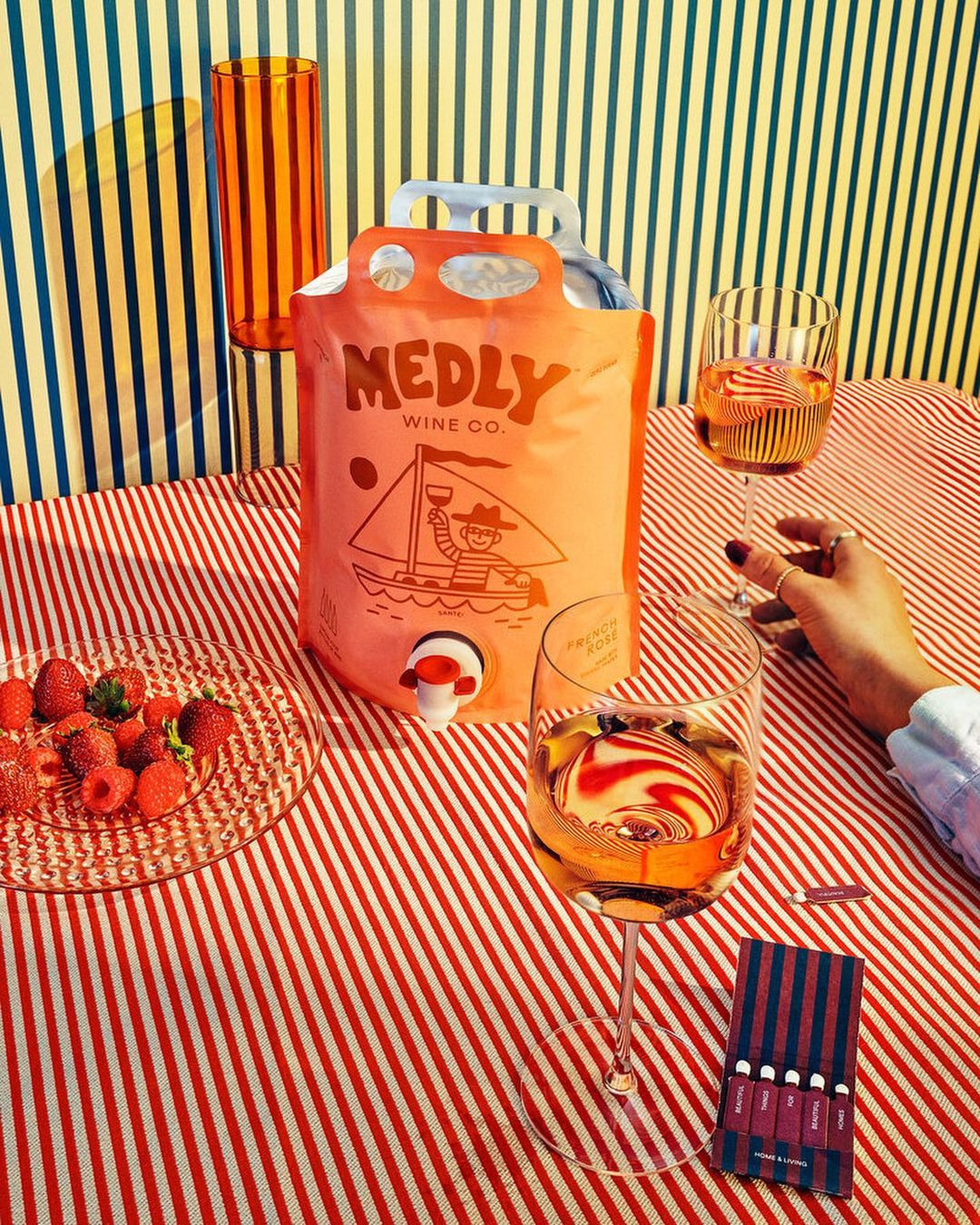 Bag-in-box wine and stemware on a striped tablecloth next to strawberries, and a matchbox with vertically striped blue and red lines.