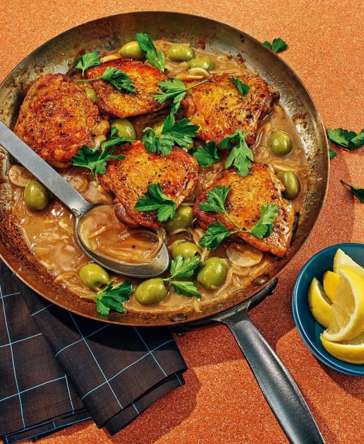 Pan-seared chicken thighs with green olives and herbs served in a copper skillet with a side of lemon wedges, creating an appetizing meal.