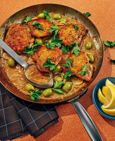 Pan-seared chicken thighs with green olives and herbs served in a copper skillet with a side of lemon wedges, creating an appetizing meal.