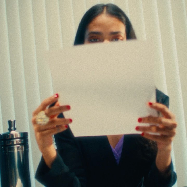 A woman with dark hair holds a blank white sheet of paper in front of her face, wearing a black blazer with red nail polish.