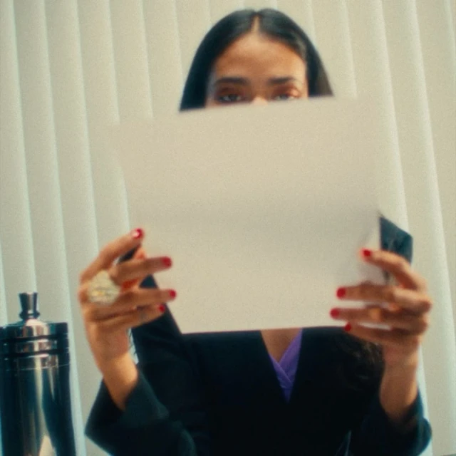 A woman with dark hair holds a blank white sheet of paper in front of her face, wearing a black blazer with red nail polish.