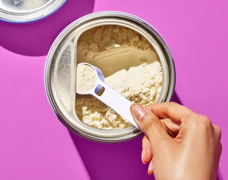 Overhead shot of a silver can with a scoop in it being held by a hand, surrounded by a bright pink background, nutritional supplement.