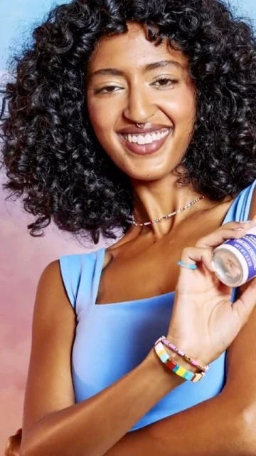 Close-up of a woman with curly dark hair and fair skin wearing a blue top and colorful jewelry, holding a product against a colorful background.
