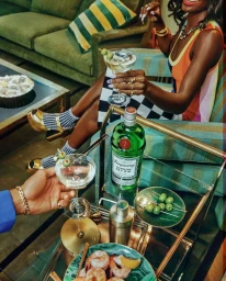 Woman holding a martini cocktail next to a bar cart with gin bottle, shrimp cocktail, and olives in a retro green living room.
