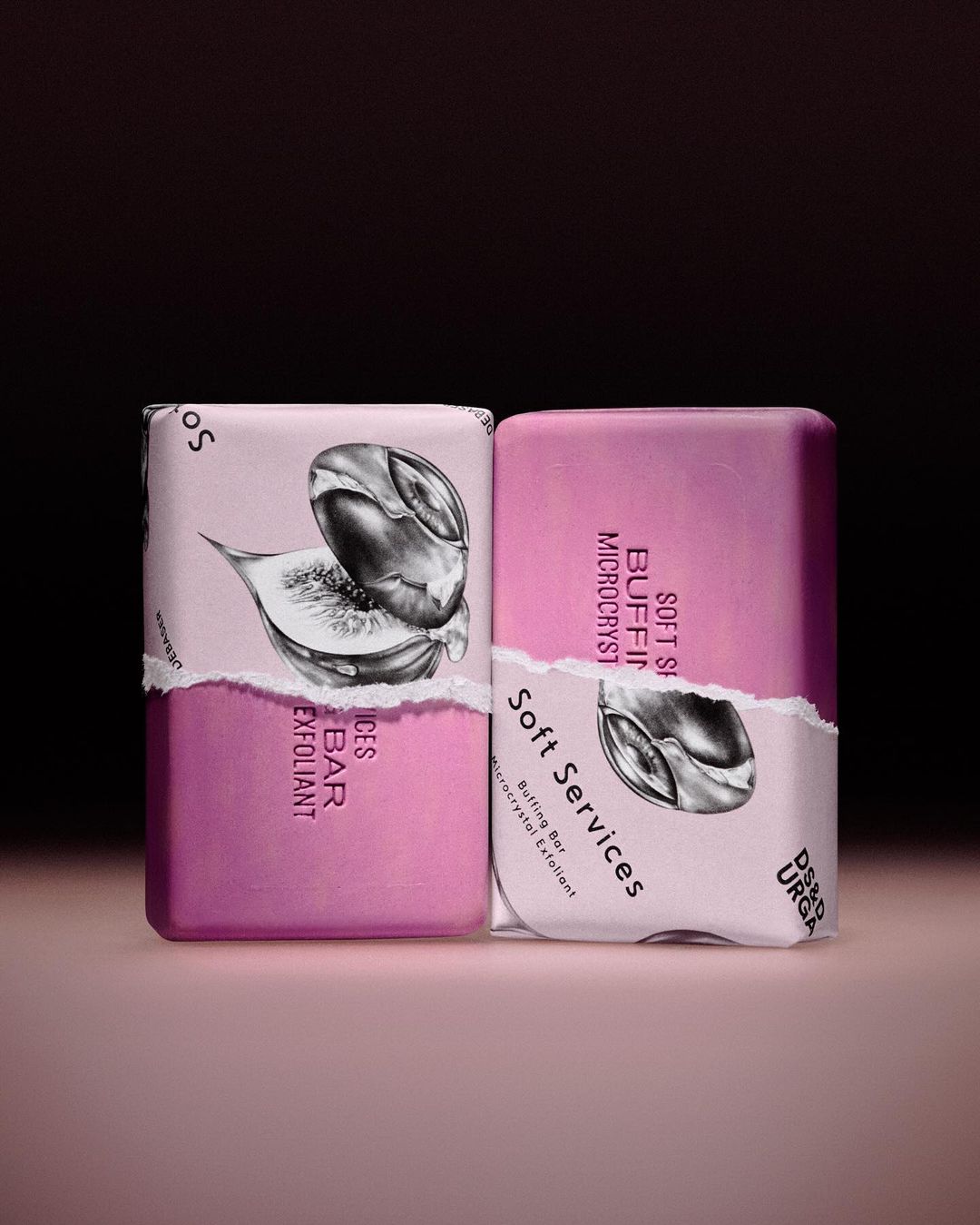 Two bars of fig-scented exfoliating soap with torn paper labels in a soft studio light against a dark mauve and gradient taupe background.