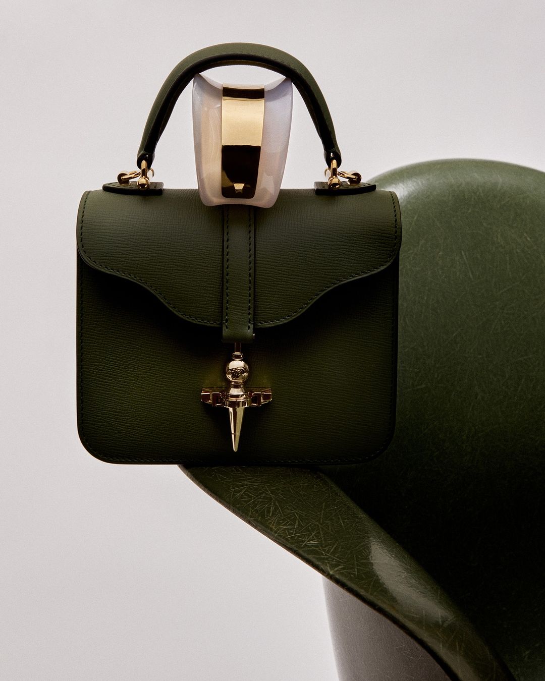 Elegant olive green leather handbag on a matching chair against a plain white backdrop, capturing a high-fashion studio aesthetic.