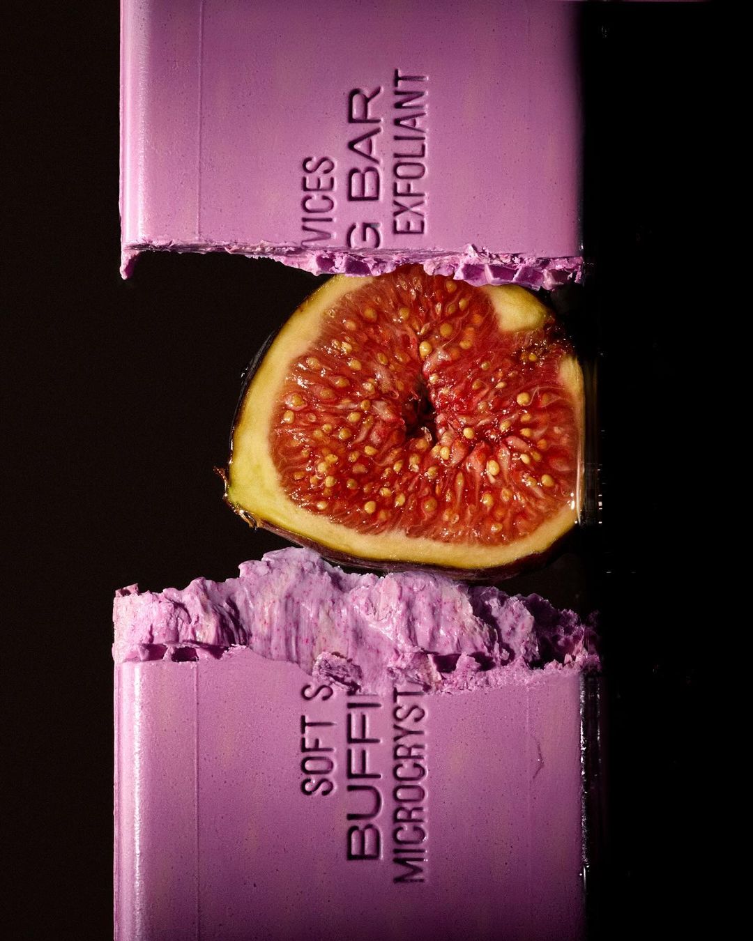 Close-up studio shot of a halved fig between two crushed bars of lilac-colored soap with embossed text against a black background.