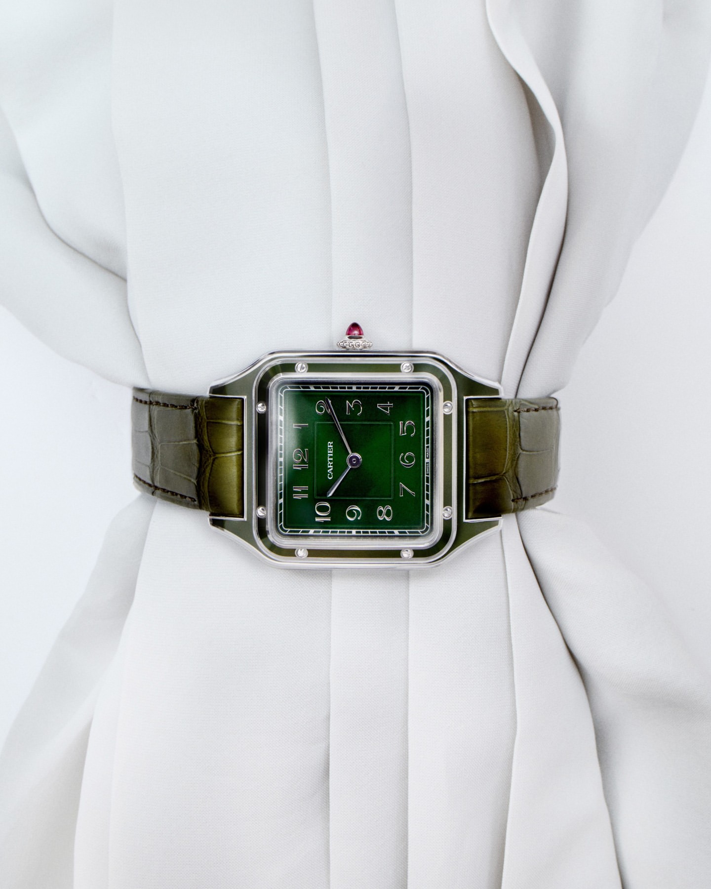 Close-up studio shot of a Cartier watch with a green face and alligator strap displayed on a white, draped garment.